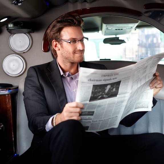 Handsome young businessman sitting in limousine, reading newspaper, smiling.